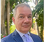 Todd Grappone in a blue suit and purple tie outdoors with trees and foliage in the background.